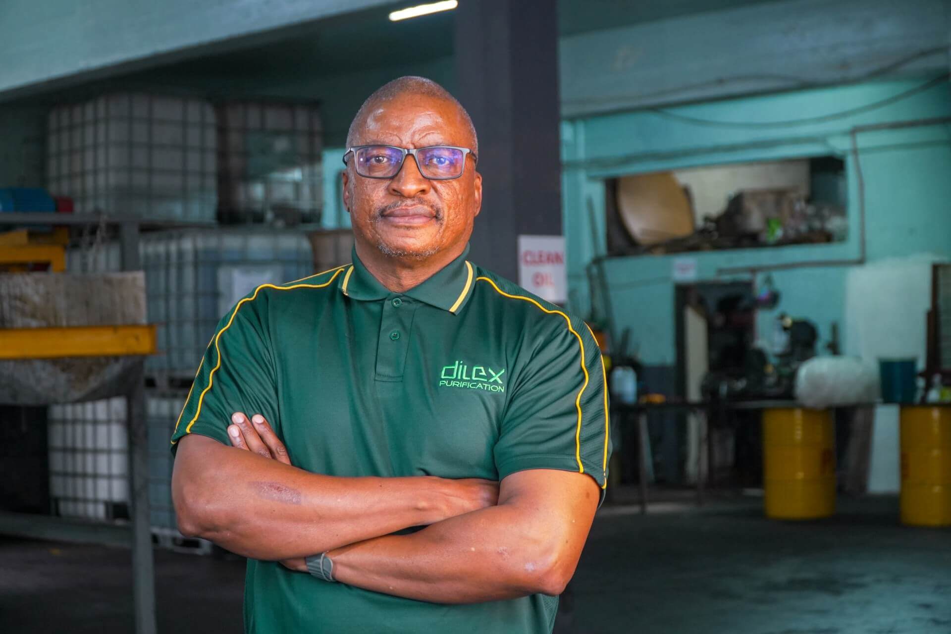 Itumeleng Rangaka, Wearing A Green Dilex Purification Shirt, Stands Confidently With Arms Crossed Inside An Industrial Oil Recycling Facility.