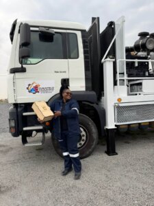Two Workers, One Wearing A Yellow Safety Vest And Helmet And The Other In Navy Overalls, Exchange A Document In Front Of A Drill Truck.