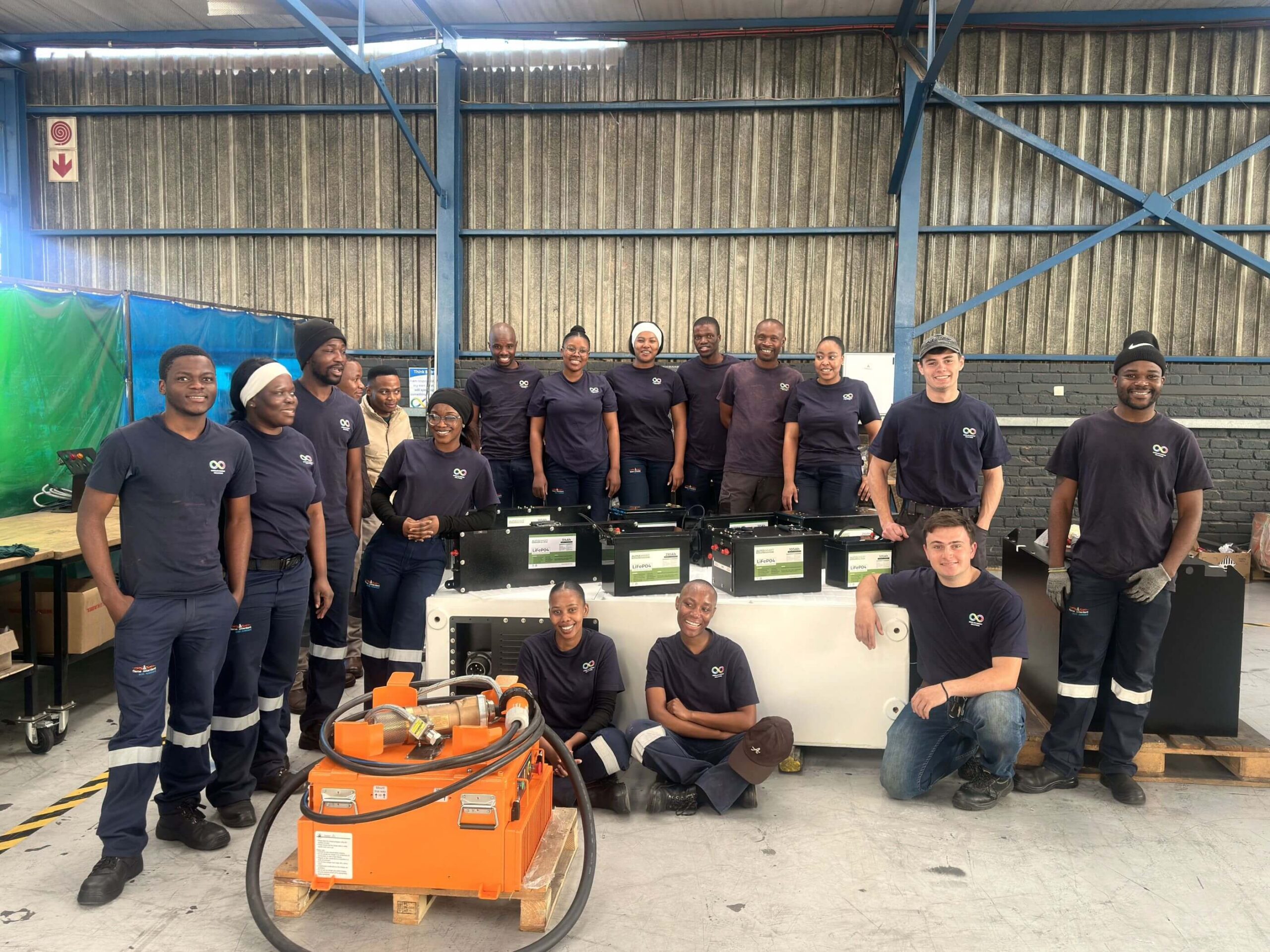 Group Photo Of Staff In Dark Coveralls In A Workshop, With Orange Equipment In Front.